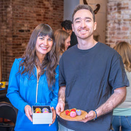 Smiling pair holding a box and wooden plate of colorful round sweets in a rustic brick-walled artisan pastry workshop.