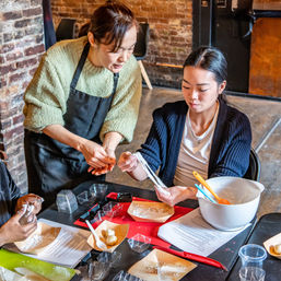 Hands-on dumpling-making class in a rustic brick-walled kitchen as an instructor guides a participant stretching dough at a table with bowls, utensils, molds and recipe sheets.