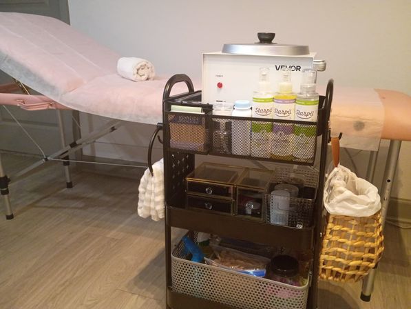 Spa treatment room with pink-covered massage table and rolled towel, metal rolling cart stocked with a wax warmer, bottles of waxing solution, creams, storage drawers and a woven waste basket.
