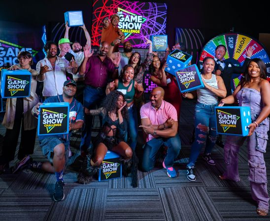 Energetic, diverse group of adults posing in a colorful indoor game show studio, holding blue challenge boxes and cheering in front of neon signage and a spinning prize wheel.