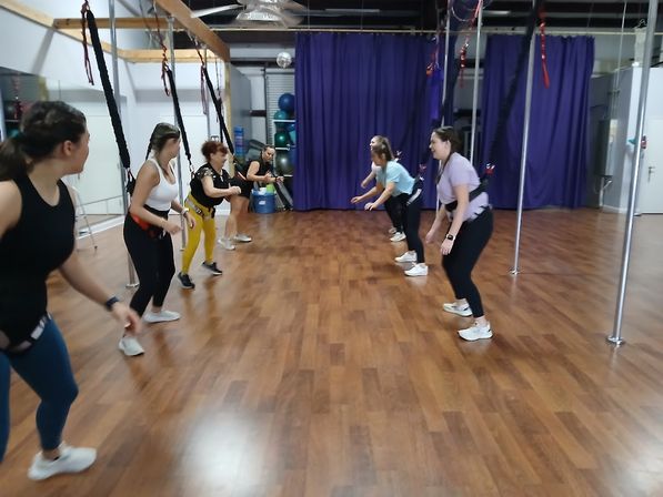 Group of women in harnesses doing a bungee fitness group class in an indoor studio with wood floor and purple curtains.