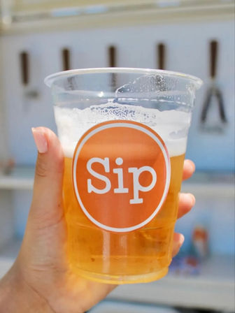 Hand holding a clear plastic cup of golden beer with a frothy head and orange circular logo, blurred outdoor event background