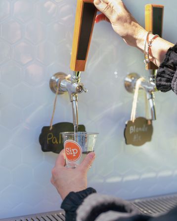 Hand pulling a wooden draft tap pouring pale ale into a clear tasting cup at a craft taproom