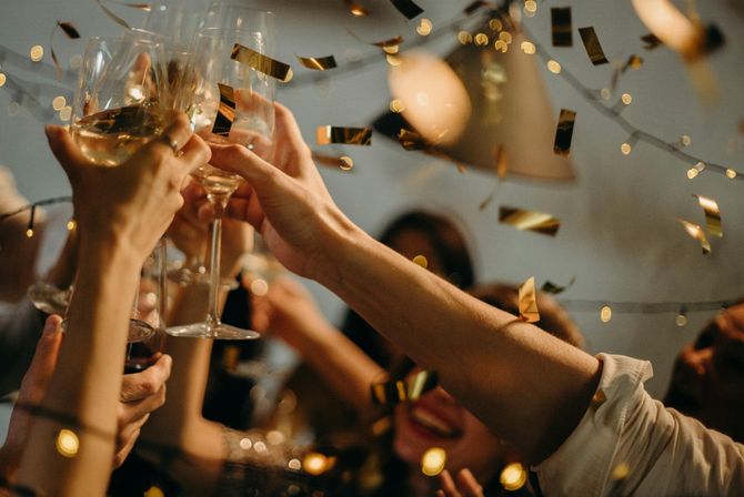 Friends clinking wine glasses amid golden confetti and twinkling lights at an indoor celebration.