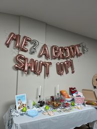 Rose-gold letter balloons reading "ME? A BRIDE? SHUT UP!" over an indoor snack table set for a bridal shower/bachelorette with candy bowls, cookies, chips, popcorn, candles and decorative pears.
