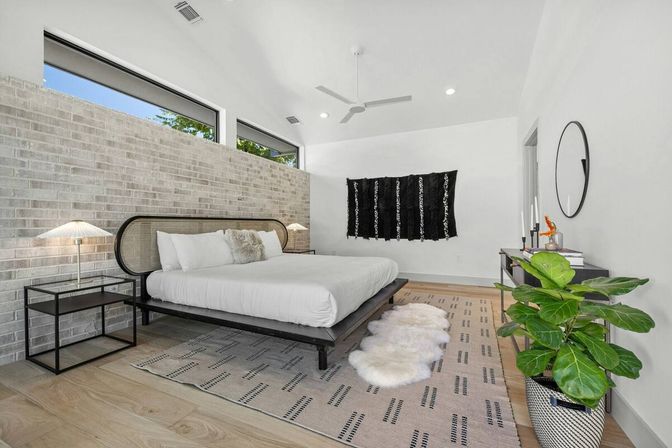 Sunlit modern minimalist bedroom with low black platform bed and white bedding against a light-gray exposed brick half-wall, clerestory windows, ceiling fan, patterned rug with faux-fur runners, black textile wall hanging, console table with round mirror and potted fiddle-leaf fig.