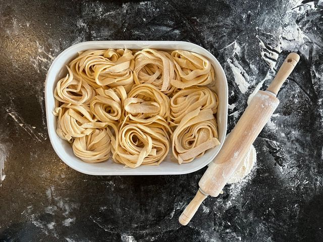 Fresh homemade fettuccine nests arranged in a white dish on a floured dark kitchen countertop beside a flour-dusted wooden rolling pin