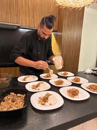 Chef adding finishing spoonfuls of creamy sauce to plated pastries topped with crumble on white plates across a wood‑paneled kitchen counter.