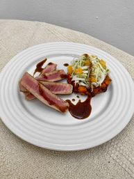 Gourmet seared ahi tuna slices drizzled with a glossy balsamic glaze, paired with citrus cabbage slaw and a roasted sweet potato stack on a white plate.