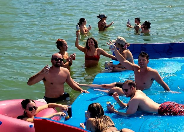 Friends at a summer lake party on bright inflatable rafts and waist‑deep water, lounging on a large blue float and a pink tube, raising canned drinks and smiling.
