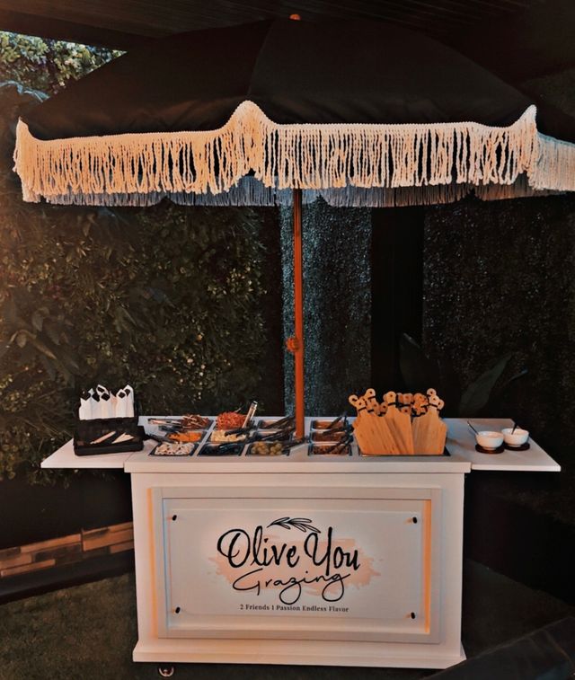 Outdoor grazing station charcuterie cart under a fringed patio umbrella in a leafy garden, with trays of olives, cheeses, wooden skewers and dipping bowls.