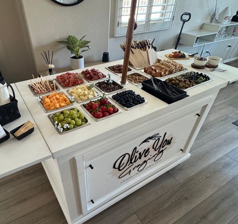 Bright modern kitchen island set up as a charcuterie buffet with trays of grapes, strawberries, blueberries, cubed cheeses, melon, cured meats, crackers, olives and serving utensils.