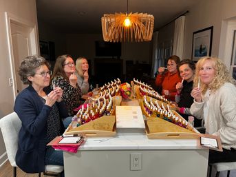 Seven women sampling scents at an at-home perfume workshop around a kitchen island, rows of colorful fragrance vials in wooden trays under a modern pendant light