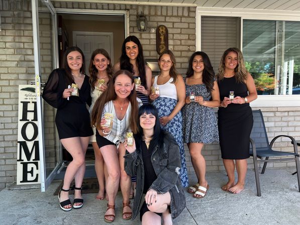Eight women smiling on a suburban brick front porch holding small gift bags, wearing casual summer outfits and sandals, with a 'HOME' welcome sign by the doorway — celebratory group photo on a sunny day.