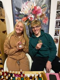 Two smiling teens seated in a fragrance boutique holding perfume bottles, colorful dropper vials on a wooden display in front of a large vibrant floral painting backdrop.