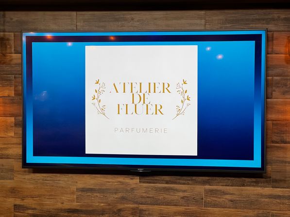 Wall-mounted digital screen on a wood-paneled wall displaying a perfume boutique logo — white square with gold floral accents and elegant gold lettering reading “Parfumerie” on a blue gradient background.