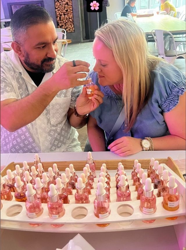 Couple sampling perfume oils at a fragrance bar — man holds a dropper bottle to a woman’s nose while a tray of pink dropper vials sits on the table during an indoor perfume tasting workshop.
