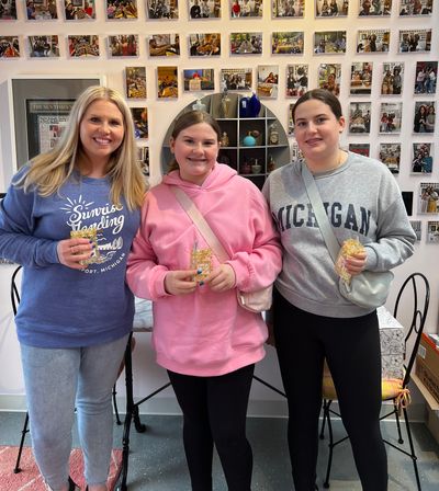 Three smiling people in casual sweatshirts (blue, pink, gray) pose inside a Michigan boutique gift shop holding small wrapped treats, with a wall of framed photos behind them.