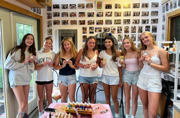 Seven smiling teens in a bright DIY perfume workshop holding small fragrance bottles, standing behind a table of colorful oils and supplies with a photo-covered studio wall in the background.