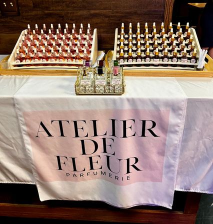 Fragrance sampling table with rows of amber and rosy dropper bottles and mini perfume vials on wooden racks over a pink-and-white tablecloth — boutique pop-up display.