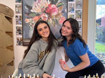 Two smiling women sampling perfume scent strips in a bright boutique with a large floral painting behind them and dropper bottles on the counter