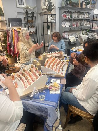 Group of women sampling artisan perfume oils at a cozy boutique fragrance workshop — tabletop trays of small dropper bottles, lit candles, snacks, and surrounding home decor shelving.
