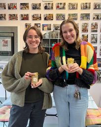 Two smiling women inside a cozy local gift shop holding small gold-wrapped gifts, one in a colorful patchwork knit cardigan, standing in front of a wall of framed customer photos.