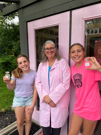 Three smiling people — a woman in a pink lab coat and two girls — posing outside a bright pink storefront door, the girls holding small perfume bottles with green shrubs in the background.