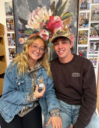 Two smiling young adults seated in a boutique; woman holds a perfume bottle in front of a large colorful floral painting and a photo-collage wall backdrop.