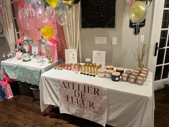 Indoor home event table display with pastel balloons, gift bags and rows of candles and fragrance bottles arranged on a white linen-covered table in front of a pink party backdrop