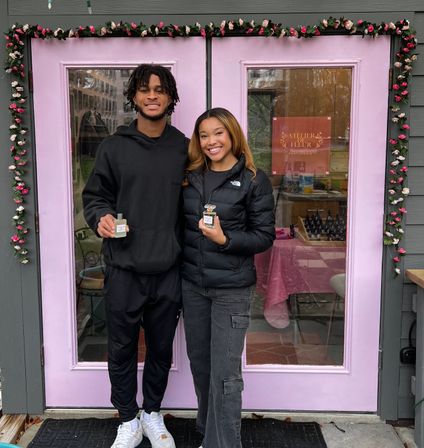Two smiling people posing at a pastel pink storefront framed with a rose garland, each holding a perfume bottle outside a cozy local perfume shop.