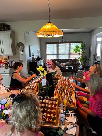 Group of women gathered around a home kitchen island sampling colorful nail polish bottles on wooden display racks, orchids on the table and a cozy living room visible in the background under a warm pendant light.