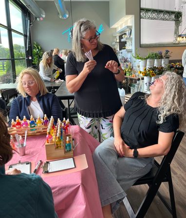 Group of women in a sunlit glass-walled floral studio at a DIY fragrance-blending workshop, instructor holding scent strips while participants sample colorful dropper bottles on a pink-covered table