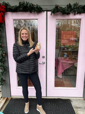Smiling woman in a black puffer jacket poses in front of pink double doors with evergreen holiday garland, holding a small gold candle or ornament outside a local boutique entrance.