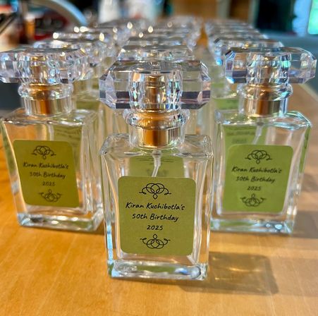 Clear mini perfume bottles with faceted crystal-like caps and green labels marked "50th Birthday 2025", neatly lined up as party favors on a wooden table