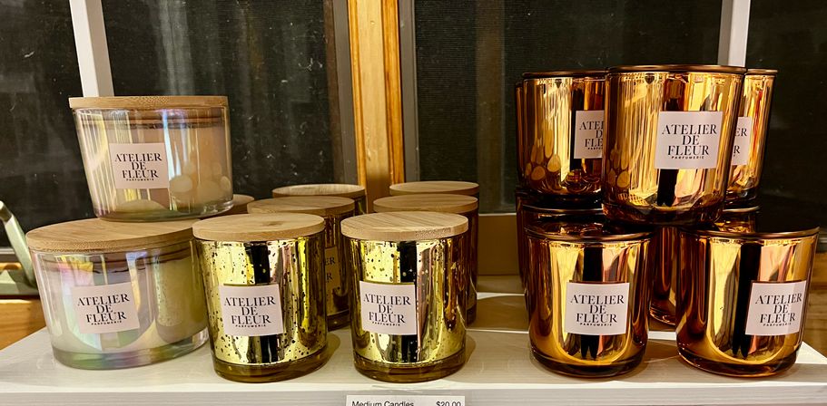 Gift-shop display of luxury scented candles in clear glass jars with wooden lids and shiny gold and copper metallic containers, neatly arranged on a white shelf.