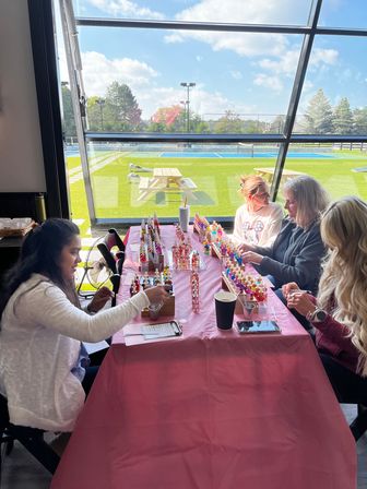 Women at a pink-covered table sampling colorful nail polish bottles during a sunny indoor workshop with large window overlooking green turf and tennis courts