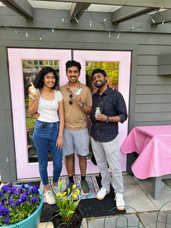 Three smiling friends pose on a front porch with pastel pink double doors holding wrapped treats, string lights overhead, purple potted flowers and a pink-covered table adding springtime vibes.