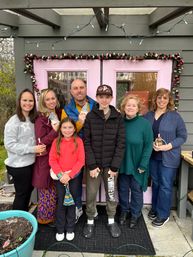 Cheerful multigenerational group of seven on a front porch in front of pink double doors framed by a floral garland and string lights, casually dressed and holding small gifts — outdoor family portrait.