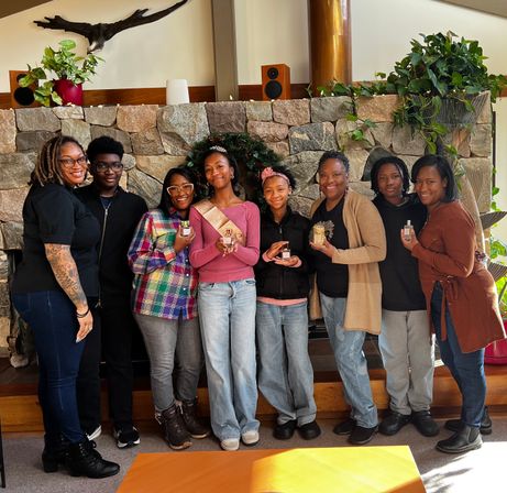 Eight-person multigenerational group smiling and posing in front of a stone fireplace with indoor plants; a celebrant wearing a sash and tiara holds a small gift while others display small bottles in a cozy lodge-style living room.