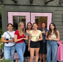 Group of six people smiling in front of a small shop with pastel pink double doors and string lights, each holding a decorated jar drink — casual outdoor summer gathering.