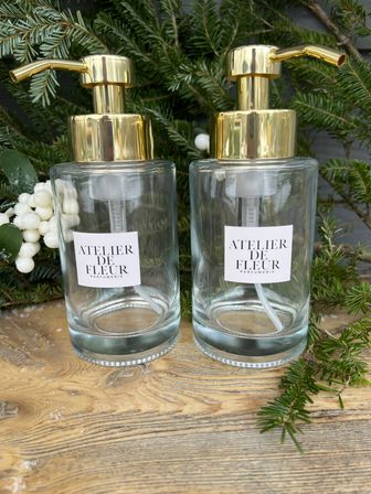 Two clear glass soap dispensers with shiny gold pumps on a wooden tabletop, surrounded by evergreen sprigs and white berry accents — chic bathroom or kitchen décor