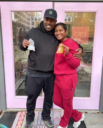 Two smiling adults in athleisure standing arm-in-arm outside a pink storefront with glass windows, holding small decorative gift bags on a patterned welcome mat.