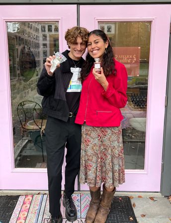 Two smiling friends pose in front of a pink boutique storefront holding perfume bottles — woman in a red jacket and paisley skirt with brown boots, man in black outfit, colorful doormat and sidewalk visible.