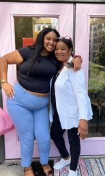 Two beaming women hugging in front of a pastel pink shop door; one in a black top and light blue pants, the other in a white cardigan and black leggings, casual street portrait