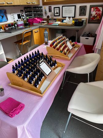 Bright beauty studio workstation with a pink-covered table holding wooden trays of blue dropper bottles and colorful polish vials, white cushioned chairs, countertops with supplies and framed wall art — tidy salon setup ready for color consultations.