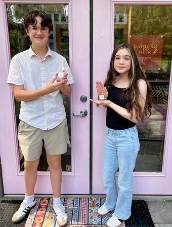 Two smiling young people standing at a pink boutique storefront door, each holding a perfume bottle over a colorful patterned welcome mat