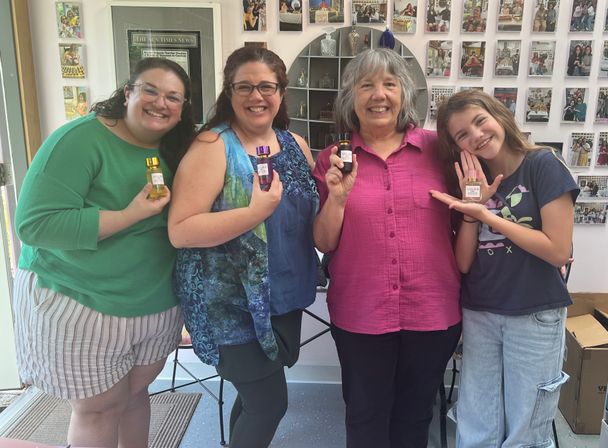 Four smiling people pose inside a bright local boutique, each holding a small bottled perfume or essential oil, with a wall of photos behind them giving an artisanal fragrance shop vibe.