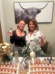 Two smiling people at a fragrance pop-up table holding perfume bottles, rows of dropper serums and scented candles, floral vase centerpiece, and a large black-and-white bull print on the wall behind them.