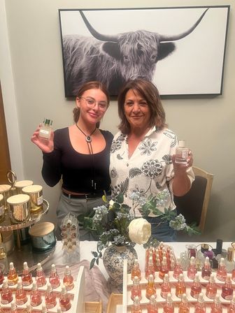 Two smiling people at a fragrance pop-up table holding perfume bottles, rows of dropper serums and scented candles, floral vase centerpiece, and a large black-and-white bull print on the wall behind them.